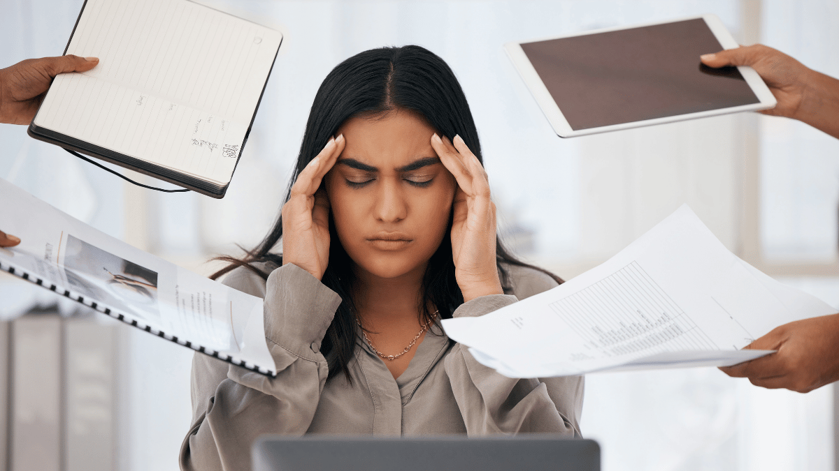 Burnout Symptoms - woman at a desk, with her eye shut, holding her head.